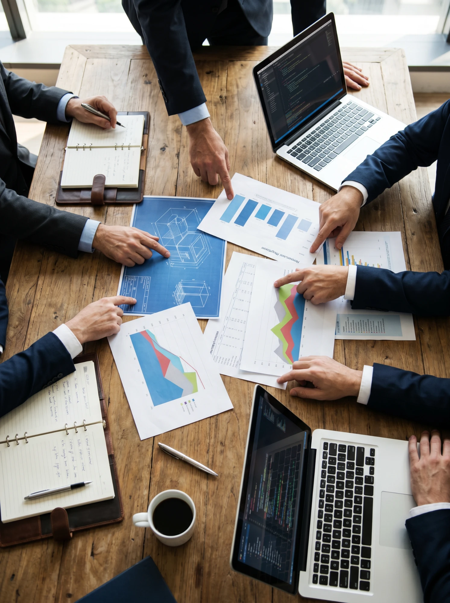Corporate team in suits reviewing data charts, blueprints and laptops around a wooden conference desk — specialty occupation professionals